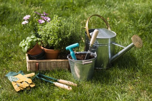 A neatly maintained front garden featuring a lush, green grassy lawn with a variety of gardening tools and supplies arranged in the foreground. There is a small wicker tray holding potted plants including flowering purple blooms and green foliage, positioned beside a metal watering can with a wooden handle. In front of the tray, garden gloves, a trowel, and pruning shears are laid out on the grass, indicating recent or ongoing gardening activity. The grass appears healthy and well-kept, with natural sunlight illuminating the scene, suggesting fair weather. The overall area is a tidy outdoor space typical of residential properties in Knightsbridge, with a focus on general gardening and lawn care that fits within the scope of services provided by Gardening Services Knightsbridge. This image captures the essential tools used for maintaining flower beds and lawn areas in a professional garden setting.