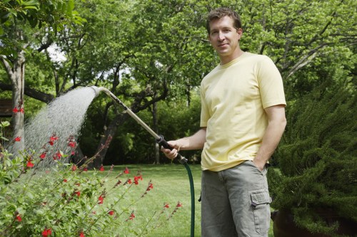 A man stands in a well-maintained garden, watering plants and flowers with a garden hose under a partly cloudy sky. The garden features a lush, green lawn with dense trees and hedges in the background, providing privacy and natural beauty. Flower beds border the lawn, filled with red and pink blooms, and are surrounded by organic soil and mulch. A paved pathway runs alongside the garden, leading towards shaded areas beneath the trees. The garden appears vibrant and healthy, with natural light highlighting the variety of greenery. The man, dressed casually in a yellow t-shirt and grey cargo shorts, is engaged in outdoor maintenance, demonstrating typical gardening activities. This scene reflects professionally cared-for outdoor spaces often serviced by companies such as Gardening Services Knightsbridge, specializing in lawn care, planting, and landscape maintenance in the local area near Knightsbridge and SW1 postcode.