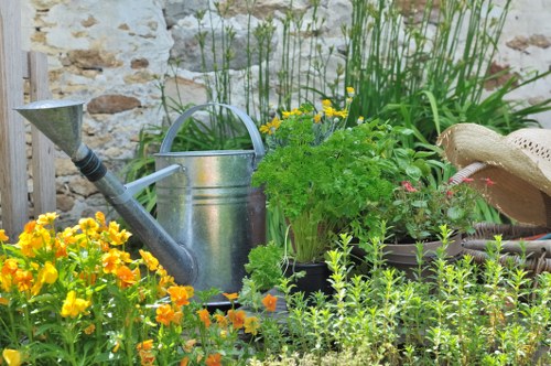 A close-up view of a small garden scene featuring a weathered stone wall in the background, with a variety of plants and garden elements arranged in front. The foreground showcases vibrant yellow and orange flowers, possibly marigolds, growing densely along the ground. Behind these, there are lush green herbs or shrubs with fine, leafy foliage, positioned next to a galvanized metal watering can with a curved handle. To the right, a woven straw hat rests on a dark container, partly obscured by tall, green grasses or reeds. The garden surface appears to be a mixture of soil and pebbles, with some elements such as the watering can and plant pots arranged casually, suggesting a well-maintained but natural outdoor space typical of a small garden in Knightsbridge or nearby areas. The scene is lit with natural daylight, capturing a clear and inviting atmosphere that highlights the diverse textures and natural tones of the garden elements, fitting within the scope of professional gardening and landscaping services offered by Gardening Services Knightsbridge.